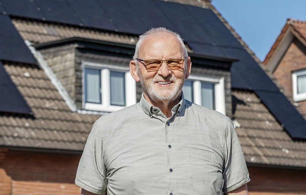 Lächelnder Mann steht vor einem Einfamilienhaus mit Solarpaneelen auf dem Dach